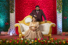 Wedding reception photography in Hyderabad by Click N Capture Studio. A bride in a stunning golden lehenga sits on an ornate golden sofa, accompanied by the groom in a maroon suit against a vibrant red floral backdrop. Contact 8143937367 for premium wedding photography services in Hyderabad.