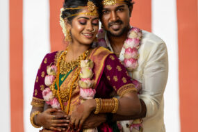 Traditional Indian wedding photography in Hyderabad by Click N Capture Studio. A bride in a vibrant green and magenta silk saree with gold jewelry and a groom in an ivory dhoti embrace lovingly, adorned with lotus garlands at a cultural wedding venue. Contact 8143937367 for professional wedding photography services in Hyderabad.