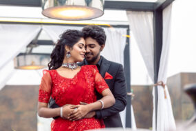 Romantic candid wedding photography in Hyderabad by Click N Capture Studio. A couple elegantly dressed, with the bride in a stunning red saree and the groom in a classic black suit, sharing a tender moment at a beautifully decorated outdoor venue. Contact 8143937367 for premium wedding photography services in Hyderabad