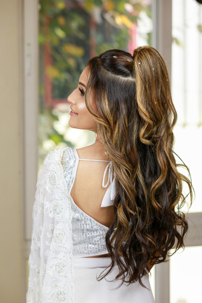 Rear view of a South Indian bride in a white saree with beautiful designs, showing her long wavy hairstyle for a reception look.